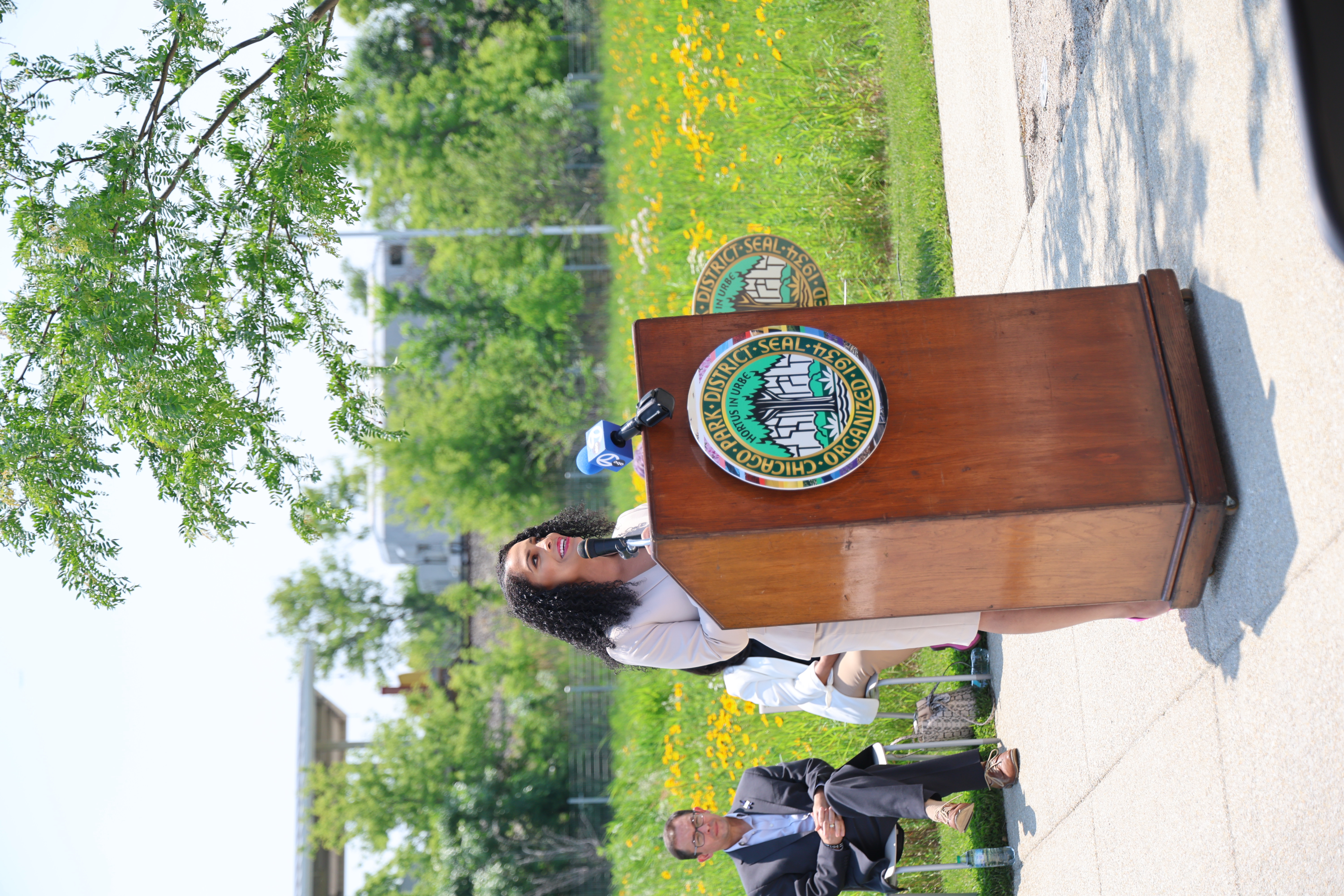 Woman speaks at podium with Chicago Park District seal. Man sits in audience. Greenery and flowers in background.